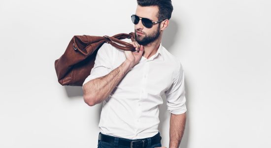 Ready to go. Handsome young man in white shirt carrying leather bag on shoulder and looking away while standing against white background